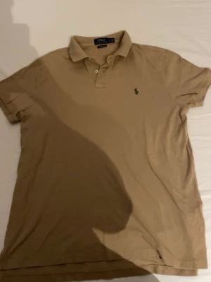 Beige pikétröja från Polo Ralph Lauren - Klassisk beige pikétröja från Polo Ralph Lauren med korta ärmar och krage. Tröjan har två knappar vid halsen och den ikoniska lilla logotypen broderad på bröstet. Tillverkad i mjuk bomull för en bekväm känsla.