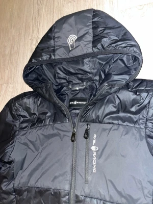 Svart dunjacka från Sail Racing - Snygg svart dunjacka från Sail Racing med huva och dragkedja framtill. Jackan har två sidofickor med dragkedja och logga på bröstet samt huvan. Perfekt för kalla vinterdagar och riktigt stilren design.