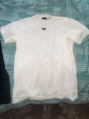 Vit t-shirt med metallogo - Säljer en stilren vit t-shirt i bomull. T-shirten har korta ärmar och en ikonisk metallogo framtill vid halsen. Enkel och clean design som passar perfekt till jeans eller shorts.