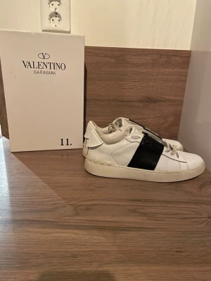 Valentino Garavani vita sneakers - Säljer ett par vita sneakers från Valentino Garavani med en bred svart rem över sidan. Skorna har klassisk låg profil, vita snören och detaljer i gummi runt sulan. Tillverkade i läder för en lyxig känsla och stilren look. Kommer med original box!