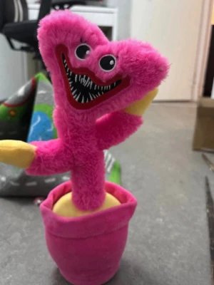 Rosa mjukisdjur monster i kruka - Huggywuggy mjukisdjur som föreställer ett monster med stort leende och spetsiga tänder. Hela figuren är gjord i mjuk plysch, har stark rosa färg och sitter i en rosa kruka med gul botten. Ögonen är broderade och uttrycksfulla. Perfekt som dekoration eller rolig present. Härmar när man pratar. 