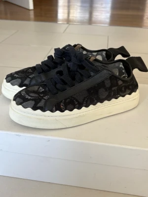 Svarta spets-sneakers från Chloé - Unika svarta sneakers från Chloé med snygga spetsdetaljer och bred vit sula. Skorna har svarta skosnören, vågig kant och stor logga bak på hälen. Materialmix av spets och läder ger en lyxig och trendig stil. Pris går att diskuteras.   