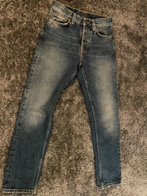 Blå tvättade jeans från Nudie Jeans - Säljer ett par blå tvättade jeans från Nudie Jeans med klassisk femficksmodell och raka ben. Jeansen har snygga slitningar och kontrastsömmar samt Nudie-logga på bakfickan. Tillverkade i kraftig denim, perfekta för en avslappnad stil.