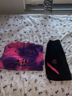 PSG träningsset lila/rosa Nike - Säljer ett fett PSG träningsset från Nike med långärmad tröja i lila och rosa med halv dragkedja och klubbmärke på bröstet. Tillhörande svarta byxor med rosa detalj och PSG-logga. Perfekt för fotbollsträning eller chill. Materialet är syntetiskt och andas.