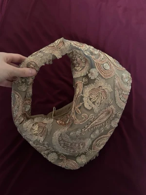 Beige paisleymönstrad handväska - Unik handväska i beige tyg med broderat paisleymönster i guld och koppar. Väskan har en mjuk, rundad form och bred axelrem. Materialet är textil med dekorativa detaljer och fransig kant. Perfekt för dig som vill sticka ut med en bohemisk och vintageinspirerad look.