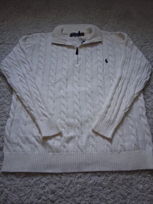 Vit kabelstickad tröja Polo Ralph Lauren - Kabelstickad vit tröja från Polo Ralph Lauren med halv dragkedja och klassisk broderad logga på bröstet. Tröjan har ribbade muddar och krage, perfekt för lager på lager och en clean look.