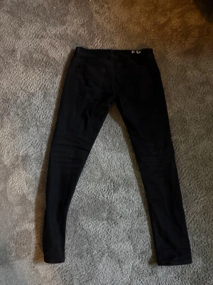 Svarta skinny jeans från Dindup - Säljer ett par svarta skinny jeans från Dindup med klassisk femficksdesign och logga på linningen. Jeansen har smal passform och är tillverkade i stretchigt denimtyg för en bekväm känsla. Perfekta för dig som gillar en clean och stilren look.