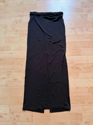 Svart långkjol med slits - En svart långkjol i mjukt och följsamt material. Kjolen har en hög midja och en snygg slits framtill som ger extra edge. Perfekt att styla med crop top eller oversized tröja för en trendig look.