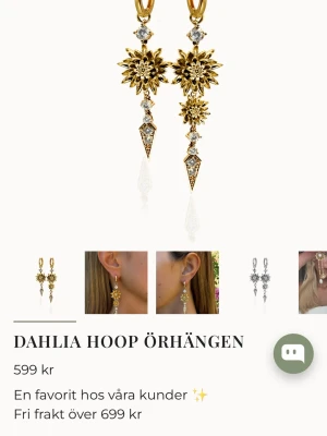 Dahlia Hoop örhängen i guld - Snygga Dahlia Hoop örhängen från Lily and Rose i guldfärgad metall med gnistrande stenar. Örhängena har en unik design med blommor och hängande detaljer som ger en lyxig känsla. Perfekta för dig som vill sticka ut med dina accessoarer.