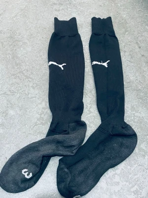 Svarta knästrumpor från Puma - Säljer ett par svarta knästrumpor från Puma med vit logga på skaftet. Strumporna är ribbade upptill och har en sportig look, perfekta till träning eller fotboll. Materialet känns som en blandning av bomull och syntet för bra komfort och passform.