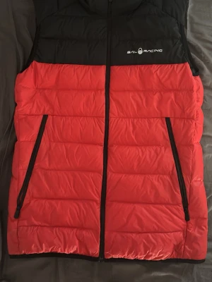 Väst sail racing - Snygg dunväst från Sail Racing i rött och svart i storlek 176 men passar de med S. jag har S/M och västen sitter fortfarande bra. Västen är i nyskick använt västen ett fåtal gånger inga defekter. Priset är inte hugget i sten. 