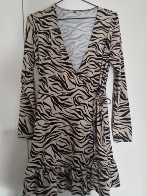 Zebramönstrad omlottklänning - Snygg kort omlottklänning med zebramönster i svart och beige. Klänningen har lång ärm, volang nertill och knyt i midjan. Djup v-ringning framtill ger en trendig look. Perfekt för dig som vill sticka ut med ett djurmönster.