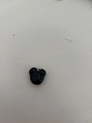 Svart berlock mussepigg - En liten svart berlock i form av ett mussepigg huvud, perfekt att använda som hänge till halsband eller armband. Tillverkad i lera med en blank yta och ett litet hål för att trä igenom kedja eller snöre.