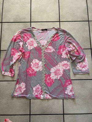 Blommig topp med spets från Mary Bell storlek L - Grå och rosa topp från Mary Bell med blommigt mönster och dekorativ spets längs v-ringningen. Toppen har trekvartsärm och är tillverkad i ett mjukt, stretchigt material. Perfekt för dig som gillar färg och detaljer!