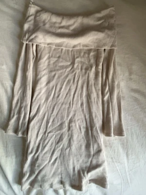 Beige ribbad offshoulder klänning - Trendig beige kortklänning med ribbad struktur och offshoulder-design. Klänningen har långa ärmar och är gjord i ett mjukt, stretchigt material som sitter snyggt på kroppen. Perfekt för dig som gillar en enkel men stilren look.