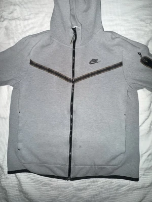 Grå Nike hoodie med dragkedja - Snygg grå hoodie från Nike med hel dragkedja framtill och svart Nike-logga på bröstet. Hoodien har svarta och mörkgrå detaljer i form av linjer över bröstet och en huva med dragsko. Perfekt för en sportig och avslappnad stil.