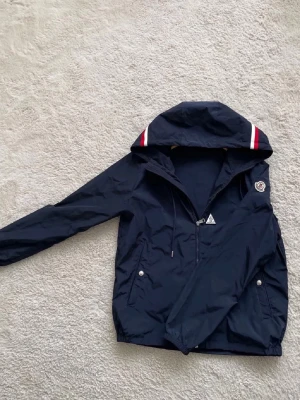 Marinblå Moncler vindjacka med huva - Snygg marinblå vindjacka från Moncler med huva och dragkedja. Jackan har vita och röda detaljer på huvan samt Moncler-logga på ärmen. Två fickor framtill med knappar. Perfekt för blåsiga dagar och stilren look och byten kan va intressant 
