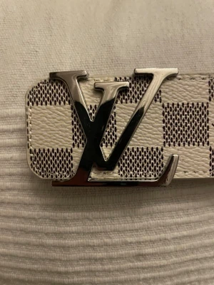 Louis Vuitton rutigt bälte i skinn - Snyggt bälte från Louis Vuitton med klassiskt rutigt mönster i vitt och grått. Bältet har en stor silverfärgad LV-logga som spänne och är gjort i skinn. Perfekt accessoar för att lyfta din outfit.