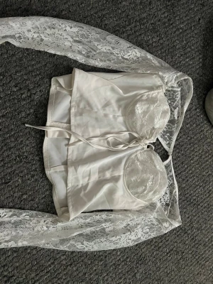 Vit spetsbody med långa ärmar - Snygg vit body med långa ärmar i spets och vadderade kupor med spetsdetaljer. Kroppen är i slät satinliknande material och har dragkedja bak. Perfekt för dig som gillar romantisk och trendig stil.
