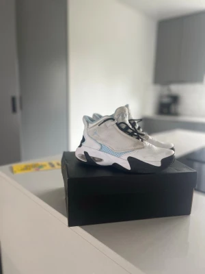 Jordan RS-2K vita och blå sneakers - Snygga Jordan RS-2K sneakers i vitt med ljusblå detaljer och svarta inslag. Skorna har en chunky sula och modern design med mesh och syntetmaterial. Perfekta för dig som gillar streetwear och vill sticka ut med en unik look.