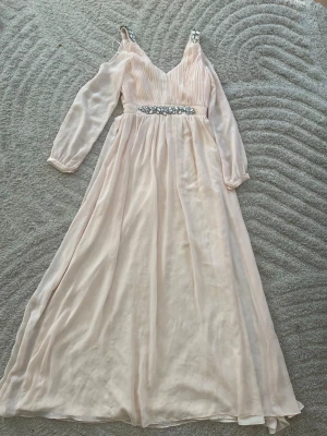 Ljusrosa långklänning med strassdetaljer - Elegant ljusrosa långklänning i chiffong med plisserad överdel och långa transparenta ärmar. Midjan pryds av en rad glittrande strassdetaljer. Klänningen har v-ringning och ett romantiskt, svepande fall.