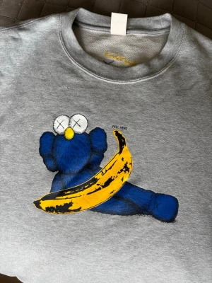 UNIQLO x KAWS x Warhol  Limited edition Grå sweatshirt  - Snygg grå sweatshirt med ett unikt tryck av en blå KAWS-inspirerad figur och en gul banan på framsidan. På baksidan finns broderad Andy Warhol-signatur och två blå kryss. Mjuk bomullskvalitet och avslappnad passform.