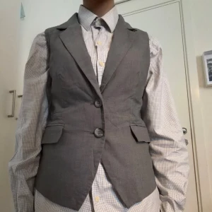 Grå kostymväst - Stilren grå väst från H&M med klassiska slag och två knappar framtill. Västen har två fickor med lock och är tillverkad i ett kostymtyg som ger en dressad look. Perfekt att bära över skjorta för en snygg och modern stil.