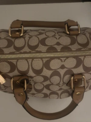 Coach beige handväska med logomönster - Snygg beige handväska från Coach med det klassiska logomönstret i brunt. Väskan har både handtag och en lång axelrem i läder, samt guldfärgade metalldetaljer. Perfekt för dig som gillar stilrena accessoarer med lyxig känsla. !inte älta!