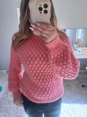 Rosa stickad tröja med bubblig struktur - Superfin rosa stickad tröja med bubblig struktur och rund hals. Tröjan har långa ärmar och ribbade muddar vid ärmslut och nederkant. Perfekt för att ge din outfit en söt och trendig touch.