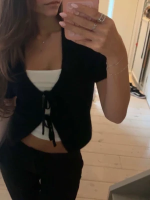 Svart kortärmad kofta från Gina Tricot - Trendig svart kortärmad kofta från Gina Tricot med knytning framtill och djup V-ringning. Koftan är stickad i ett mjukt material och har en croppad passform som passar perfekt till linne eller topp under.