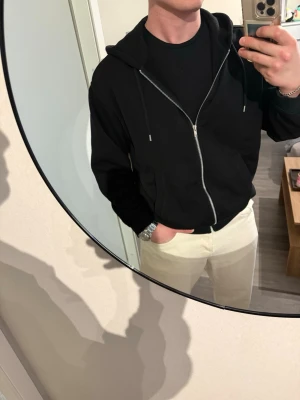Svart zip hoodie från H&M - Svart hoodie med dragkedja från H&M. Klassisk modell med huva och dragsnören, två fickor framtill och ribbade muddar vid ärmslut och nederkant. Perfekt för en avslappnad och stilren look.