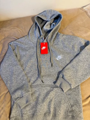Grå Nike hoodie med huva - Säljer en grå hoodie från Nike med broderad logga på bröstet och klassisk huva med dragsnören. Tröjan har ribbade muddar och en stor magficka. Materialet är mjukt och bekvämt, perfekt för chill dagar. Taggen sitter kvar.