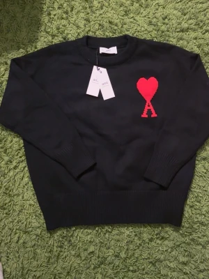 Svart stickad tröja från Ami Paris - Svart stickad tröja från Ami Paris med röd hjärtlogo och bokstaven A på bröstet. Tröjan har rund halsringning, ribbstickade muddar och är tillverkad i mjukt material. Perfekt för dig som gillar stilrena plagg med en twist.