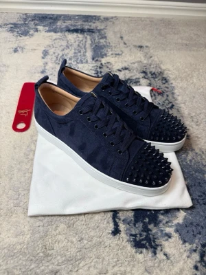 Mörkblå Louboutin sneakers med nitar - Snygga mörkblå sneakers från Christian Louboutin med matta nitar på tån och klassisk röd sula. Skorna har en låg profil, snörning och är tillverkade i textil med läderdetaljer. Perfekta för dig som vill sticka ut med en edgy och exklusiv stil.