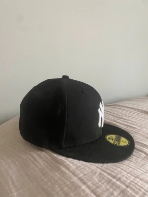 Svart New Era Yankees keps Baseball Keps One-Zize - Storlek passar perfekt för en 10-14 åring. Säljer en svart New Era 59Fifty keps med New York Yankees-logga i vitt framtill och broderad MLB-logga bak. Klassisk flat brim och broderad New Era-logga på sidan. Tillverkad i bomull och polyester, perfekt för dig som gillar streetwear och sportig stil.