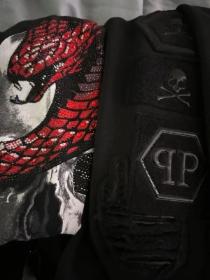 Philipp Plein Tröja och Byxor - Cool svart tröja från Philipp Plein med detaljerad röd orm i paljetter och broderad dödskalle. Tröjan har även PP-logga och unika texturdetaljer. Perfekt för dig som gillar statement-plagg och streetstyle.