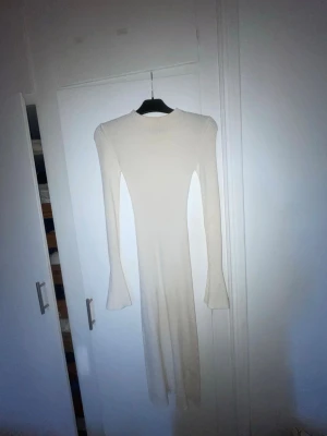 Vit ribbad långklänning Gina Tricot - Lång, vit ribbad klänning från Gina Tricot med långärmad design och lätt utsvängda ärmslut. Klänningen har en kroppsnära passform och är tillverkad i ett mjukt stickat material som ger en snygg siluett.