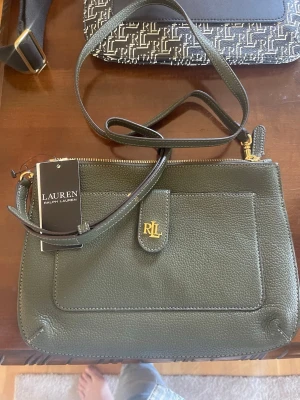 Grön handväska från Lauren Ralph Lauren - Stilren grön handväska i läder från Lauren Ralph Lauren med justerbar axelrem och guldfärgad RLL-detalj framtill. Väskan har en praktisk ytterficka och stängs med dragkedja. Perfekt för dig som gillar klassisk och enkel design.