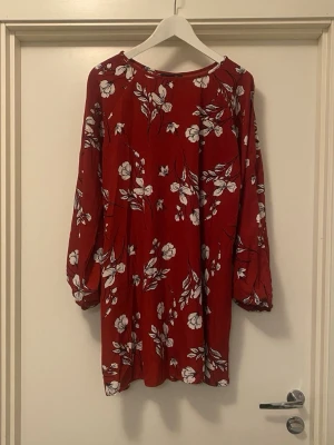 Röd blommig klänning med långa ärmar - Röd kortklänning med vitt blommönster och långa ärmar. Klänningen har en avslappnad passform och är perfekt för dig som gillar färg och mönster. Materialet känns lätt och luftigt, vilket gör den skön att bära under varmare dagar. Ärmarna har slits/öppning på sidan och vid axlarna.