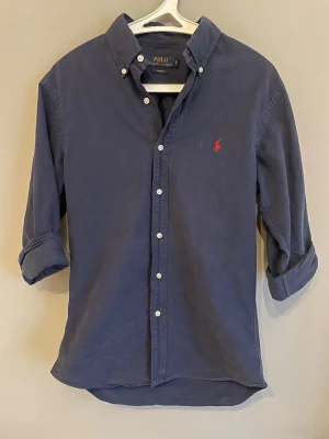Blå skjorta från Polo Ralph Lauren - Klassisk marinblå skjorta från Polo Ralph Lauren i slim fit-modell. Skjortan har långa ärmar, knappar framtill och en röd broderad logga på bröstet. Perfekt för en stilren och avslappnad look.