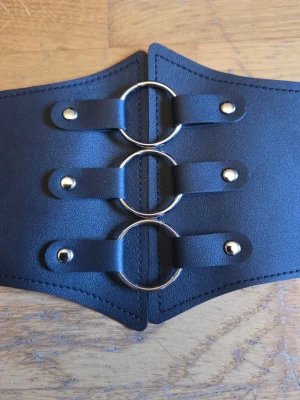 Svart midjebälte med o-ringar goth gothic alternative - Nytt oanvänt svart bälte i konstläder med silvriga metalldetaljer. Längd som minst: 82 cm. Elastiskt. Knäpps baktill med tre tryckknappar. 