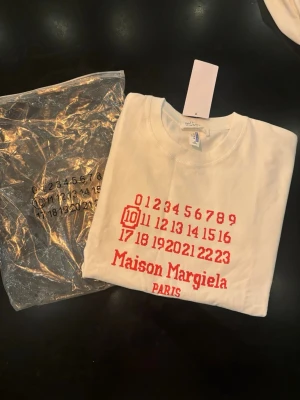  Maison Margiela t- shirt  - Vit t-shirt från Maison Margiela med rött tryck framtill som visar märkets ikoniska siffror och logga. Klassisk rund hals och korta ärmar. Perfekt för dig som gillar stilrena och trendiga plagg med designer-touch.