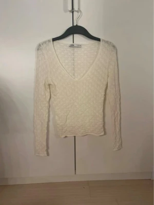 Vit spetsblus med V-ringning från Zara - Säljer en vit, långärmad spetsblus från Zara med djup V-ringning. Blusen har ett elegant mönster i spets och är tunn och lätt, perfekt för att skapa en stilren look. Materialet är mjukt och följsamt, vilket gör den bekväm att bära.