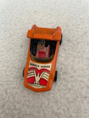 Wonder Woman leksaksbil från 1979 - Säljer en orange leksaksbil med Wonder Woman-tema från DC Comics, år 1979. Bilen har svarta hjul, detaljerad Wonder Woman-figur i förarsätet och coola dekaler med logotypen på motorhuven. Perfekt för samlare eller fans av superhjältar.