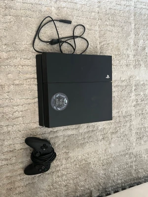 PlayStation 4 med handkontroll och kabel - Säljer en svart PlayStation 4 från Sony med en tillhörande svart handkontroll och strömkabel. Konsolen har en stilren, rektangulär design i plast och en dekorativ sticker på ovansidan. Perfekt för gaming och underhållning hemma.