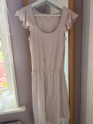 Ljusbeige klänning med volangärmar - Supersöt ljusbeige klänning med volangärmar och rund halsringning. Klänningen har resår i midjan för en bekväm passform och är tillverkad i mjukt bomullsmaterial. Perfekt för varma dagar och enkel att styla.