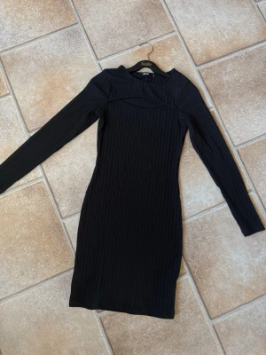Svart ribbad långärmad klänning Bik Bok - Snygg svart ribbad klänning från Bik Bok med lång ärm och en cool cut out-detalj vid bröstet. Klänningen är kroppsnära och har en kort längd, perfekt för dig som gillar en stilren och trendig look.