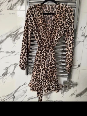 Leopardmönstrad omlottklänning med knyt - Säljer en leopardmönstrad kort omlottklänning med långa ärmar och knytband i midjan. Klänningen har en djup v-ringning. Inga skador eller defekter. 