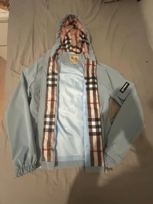 Ljusblå vindjacka med rutigt foder storlek S - Snygg ljusblå vindjacka från Burberry med klassiskt rutigt foder i huva och längs dragkedjan. Jackan har huva, resår vid ärmslut och en patch på ena ärmen. Perfekt för dig som vill ha en stilren och ikonisk look.