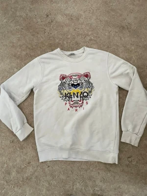 Vit Kenzo sweatshirt med tigertryck - Säljer en vit sweatshirt från Kenzo med det ikoniska broderade tigerhuvudet i flera färger på bröstet och texten 'KENZO PARIS'. Tröjan har rund halsringning, långa ärmar och ribbade muddar. Perfekt för dig som vill ha en statement piece i garderoben.